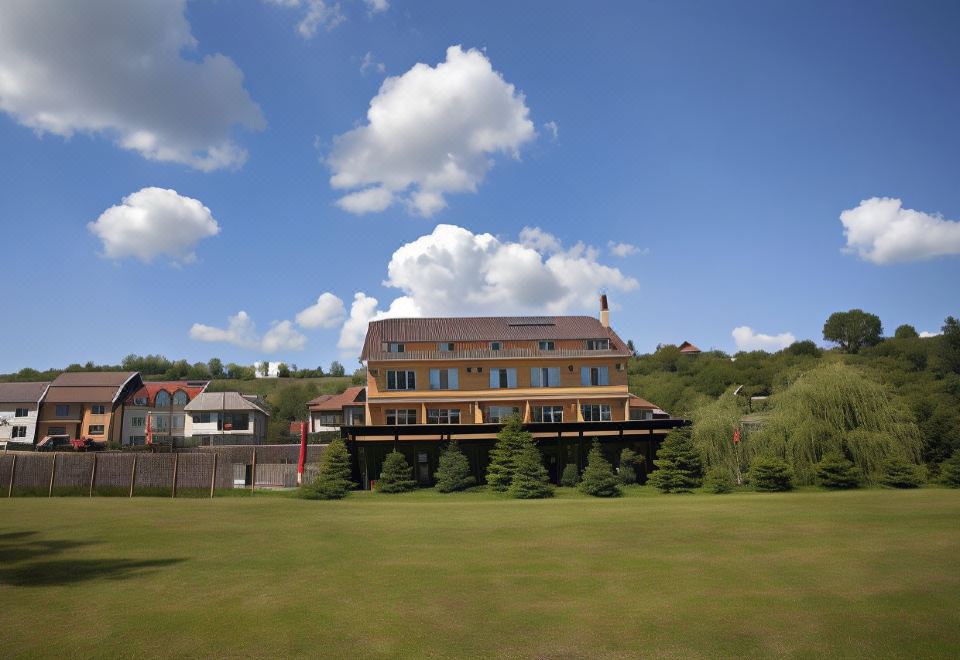 a large , brown - roofed building with a red roof and balcony , surrounded by green grass and trees , under a blue sky dotted with at Hotel Terra