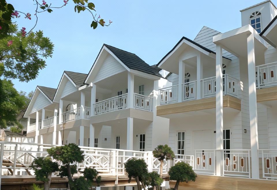 a row of white , two - story houses with balconies and railings , surrounded by green trees and clear skies at Chevilly Resort & Camp