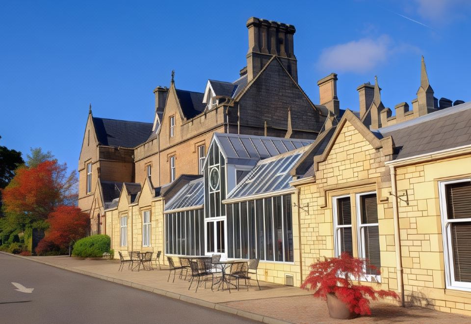 a large building with a glass - enclosed atrium and a table and chairs outside , surrounded by trees at Macdonald Inchyra Hotel and Spa