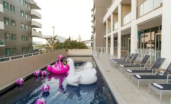 a large swimming pool with a pink flamingo and swan floaties in the middle , surrounded by lounge chairs and a building at Sage Hotel Wollongong