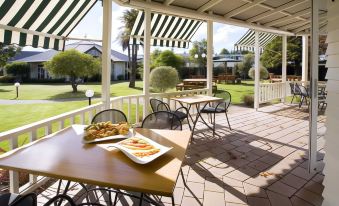 a patio with wooden tables and chairs , surrounded by green grass and trees , under a striped awning at Wairakei Resort Taupo