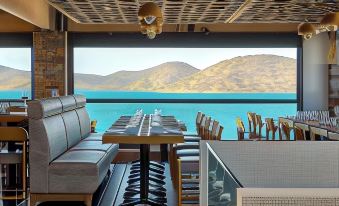 a restaurant with a view of the ocean , featuring a long dining table and several chairs at Elounda Orama