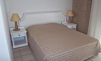 a neatly made bed with a beige quilt is flanked by two nightstands and lamps on either side at Milos Apartments