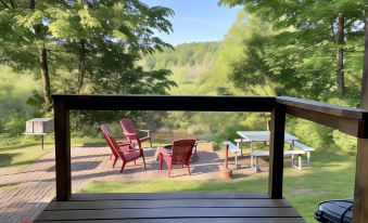 a wooden deck with three red chairs , a table , and a chair under an umbrella , overlooking a serene lake surrounded by trees at Betsie Riverside Resort