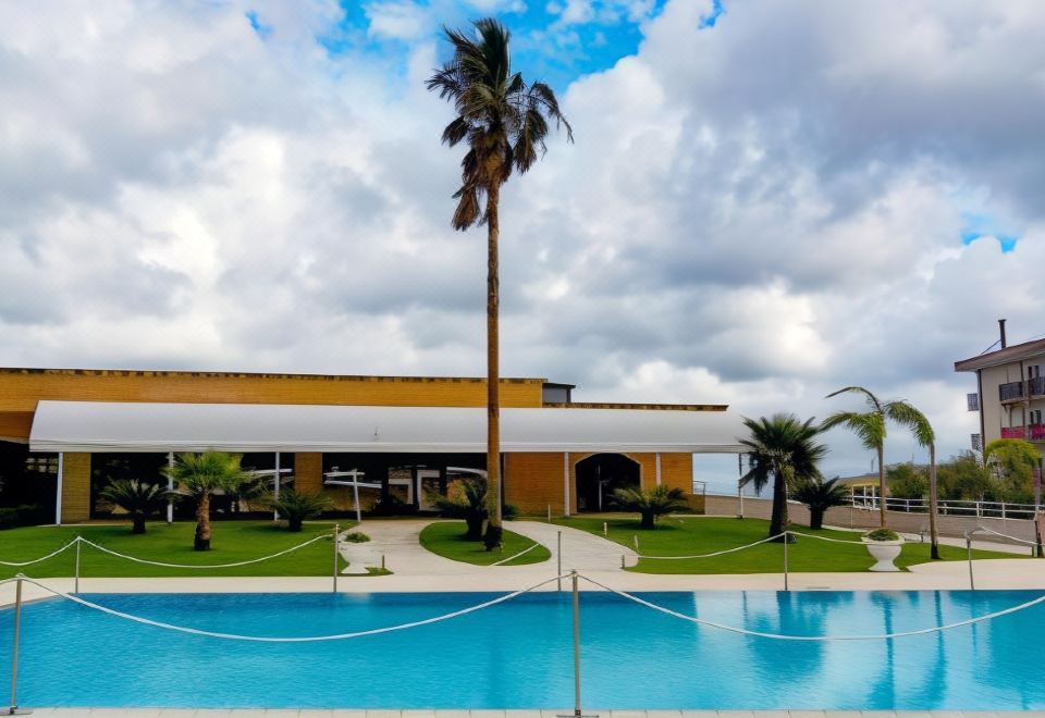 a large swimming pool surrounded by palm trees , with a building in the background and clouds in the sky at Hotel Capital