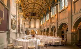 a large , elegant banquet hall with white tables and chairs , chandeliers , and arched ceilings , set for an elegant event at De Vere Beaumont Estate
