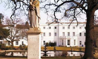 a statue of a woman is on a pedestal in front of a white building with trees and a pond at De Vere Beaumont Estate