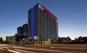 a tall building with a red and blue sign is lit up at night , with cars driving by in the foreground at Aloft Perth