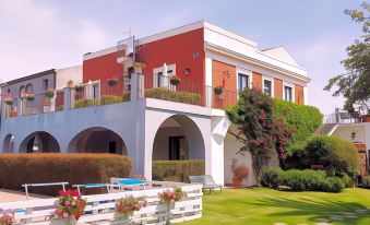 a large , red - roofed building with white columns and balconies , surrounded by lush greenery and flowers , under a clear blue sky at Etna Hotel