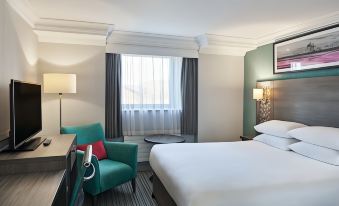 a modern hotel room with a white bed , blue armchair , and desk , along with a window offering a view of trees at Leonardo Hotel Middlesbrough