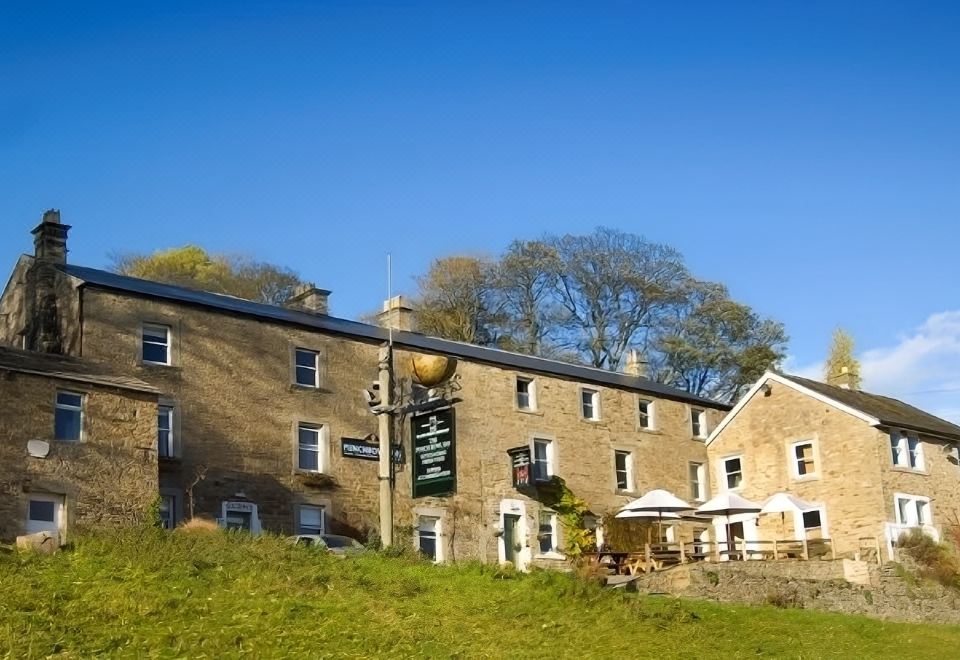 a stone building with a green sign on the side , surrounded by trees and grass at The Punch Bowl Inn