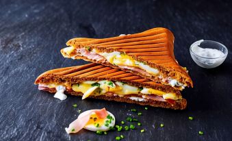 a sandwich cut in half , topped with ham , cheese , and green onions , sitting on a black plate at Ibis Longwy Mexy