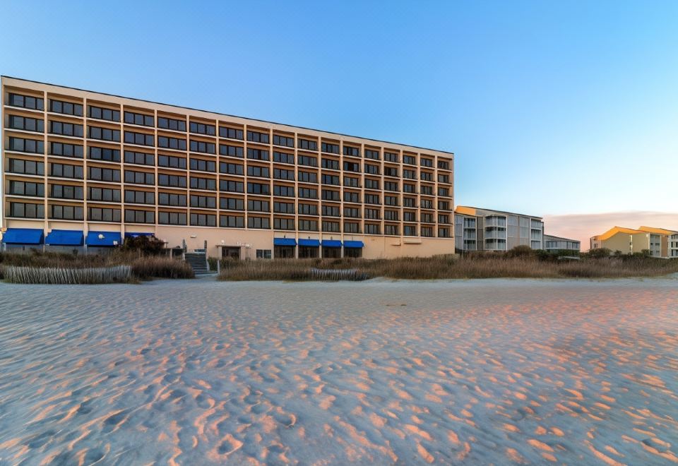 a large building with many windows is situated on the beach , and a few small buildings are visible in the background at Crystal Coast Oceanfront Hotel