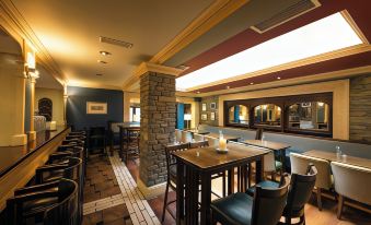 a modern bar with wooden tables and chairs , stone walls , and large windows , under a blue ceiling at Castle Hotel Macroom