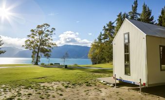 a small white house is situated on a grassy field near a lake , with mountains in the background at The Camp - Lake Hawea
