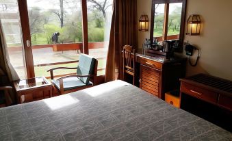 a bedroom with a bed , nightstands , and chairs is shown with a view of the outside through large windows at Seronera Wildlife Lodge