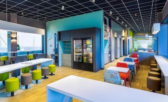 a modern cafeteria with colorful tables and chairs , a refrigerated display case , and black ceiling beams at Ibis Styles Genève Palexpo Aéroport