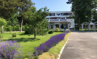 "a white building with the sign "" academy hotel "" on top , surrounded by lush greenery and purple flowers" at Academy Hotel