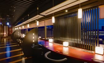 a modern bar with a long wooden table and chairs , surrounded by glass windows and lit by hanging lights at Hotel Metropolitan Akita