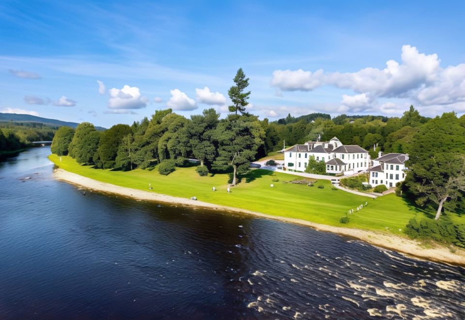 a large white building is situated on the edge of a river , surrounded by trees at Banchory Lodge Hotel
