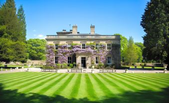 a large , stone house surrounded by lush green grass and trees , with a blue sky above at Eshott Hall