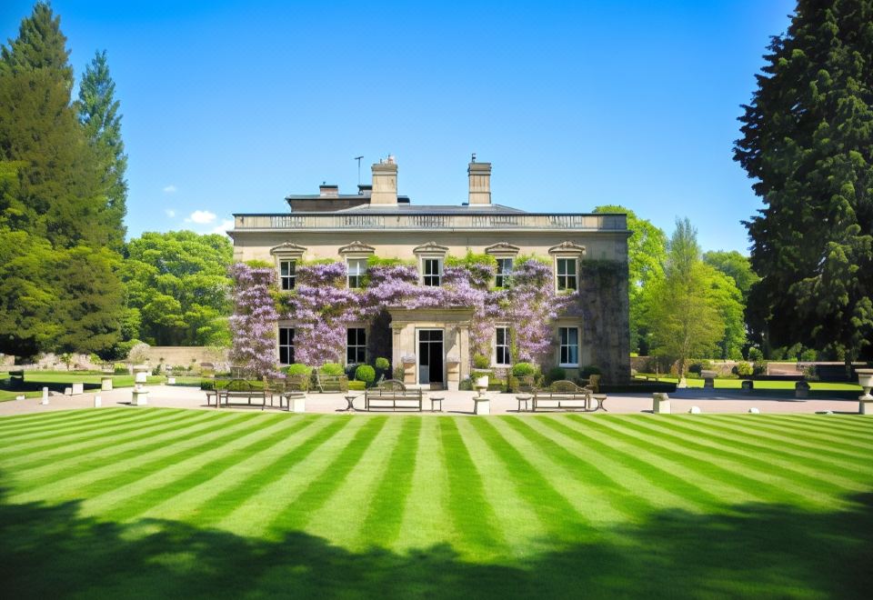 a large , stone house surrounded by lush green grass and trees , with a blue sky above at Eshott Hall