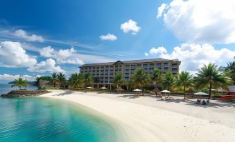 a large hotel building situated on a sandy beach , surrounded by palm trees and a body of water at Palau Hotel