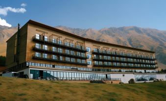 a large hotel building situated in a grassy field , surrounded by mountains in the background at Rooms Hotel Kazbegi