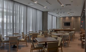 a large , empty conference room with wooden tables and chairs , white curtains on the windows , and pendant lights hanging from the ceiling at JL Hotel by Bourbon