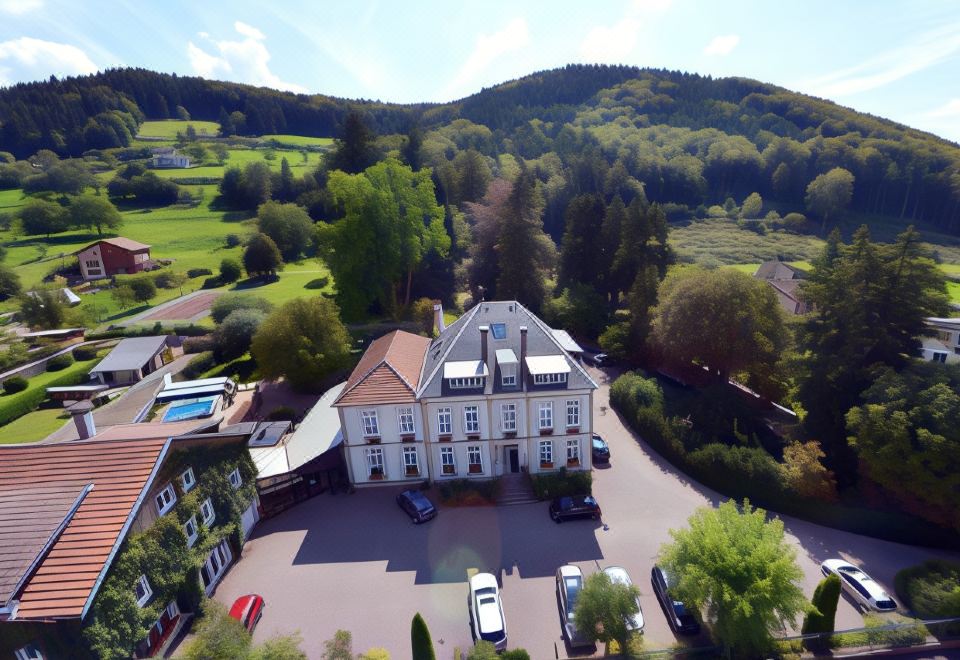 aerial view of a large white building surrounded by green trees and mountains , with cars parked in the driveway at Logis la Résidence