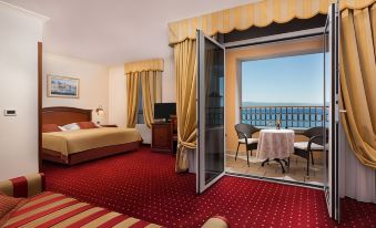 a hotel room with a red carpet and a balcony overlooking the ocean , featuring a bed , television , and balcony table at Hotel Savoy