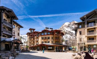 a snow - covered town square with a group of people standing in front of a building , enjoying the winter scenery at The Village at Palisades Tahoe