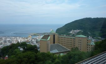 a large building with a green roof is perched on a hillside overlooking the ocean at Kamenoi Hotel Atami Annex