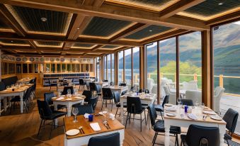 a restaurant with wooden tables and chairs , a large window offering a view of mountains , and blue placemat - covered plates on the tables at The Whispering Pine Lodge