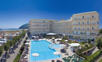 a large , inviting swimming pool is surrounded by umbrellas and lounge chairs , with a beachfront hotel in the background at Hotel Miramare