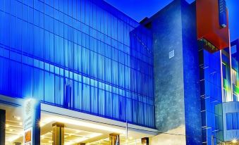 a modern building with a blue glass facade and a large open area in front at Golden Flower by Kagum Hotels