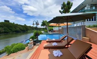 a poolside area with lounge chairs and an umbrella , overlooking a body of water under a cloudy sky at Princess River Kwai Hotel