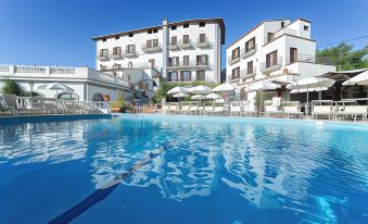 a large outdoor swimming pool surrounded by white buildings , with lounge chairs and umbrellas placed around the pool area at Hotel Jaccarino