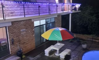 a backyard with a swimming pool , a dining table , chairs , and a rainbow umbrella , illuminated by blue lights at Hidden Inn