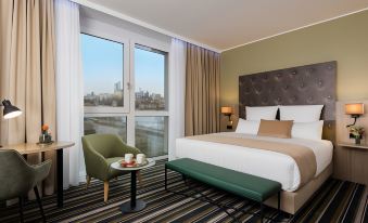 a hotel room with a large window offering a view of the city , a comfortable bed , and a green bench at Leonardo Offenbach Frankfurt