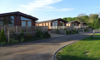 a row of wooden houses with a winding driveway and greenery in the foreground , under a blue sky at King's Lynn Caravan & Camping Park