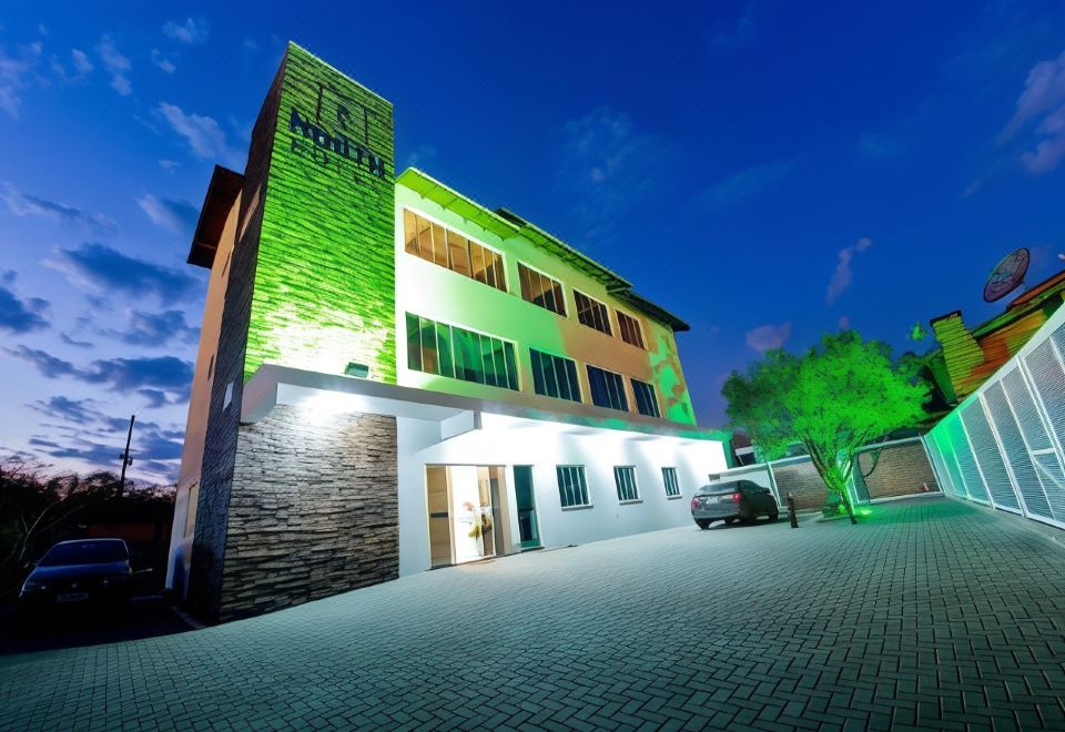 a large , modern building with green and white exterior walls , lit up at night , and a car parked outside , under a blue sky with at North Hotel - Aeroporto