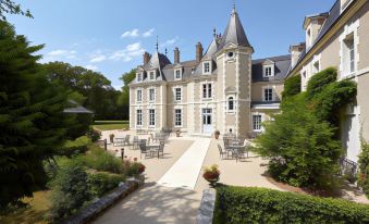 a large , two - story french chateau with a courtyard surrounded by chairs and tables , all set against the backdrop of lush greenery at Les Sources de Cheverny