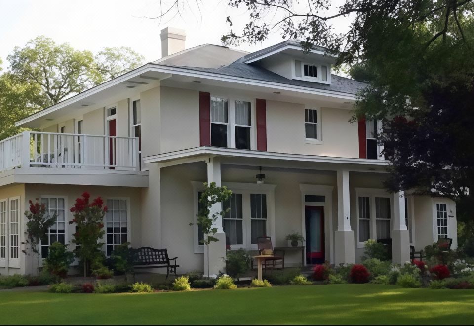 a white two - story house with red shutters , surrounded by lush greenery and flowers , and a black bench in the front yard at Delta Street Inn