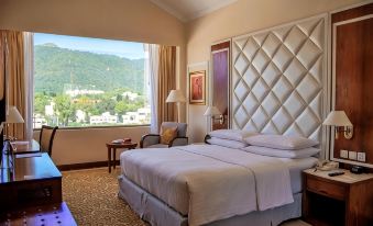 a well - lit hotel room with a large bed , white curtains , and a view of the mountains outside the window at Islamabad Marriott Hotel