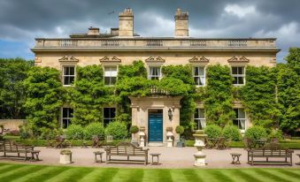 a large , lush green house with multiple windows and doors , surrounded by trees and greenery , under a cloudy sky at Eshott Hall