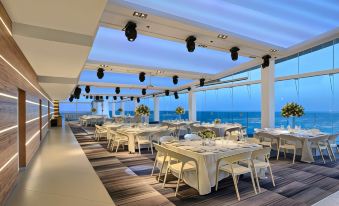 a large , modern restaurant with white tables and chairs is set up on a rooftop overlooking the ocean at Resort Hadera by Jacob Hotels