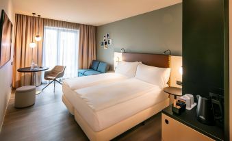 a large bed with white sheets is in a room with a blue chair and a window at Hilton Garden Inn Mannheim