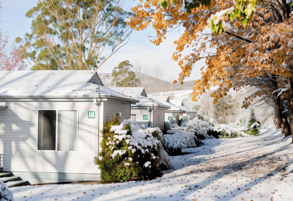 a row of white cottages are lined up on a snow - covered road , surrounded by trees at Discovery Parks - Jindabyne