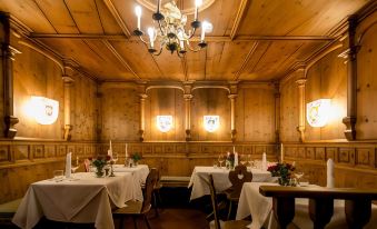 a dimly lit wooden restaurant with multiple tables and chairs , each table having its own white tablecloth and flowers at Hotel Sailer